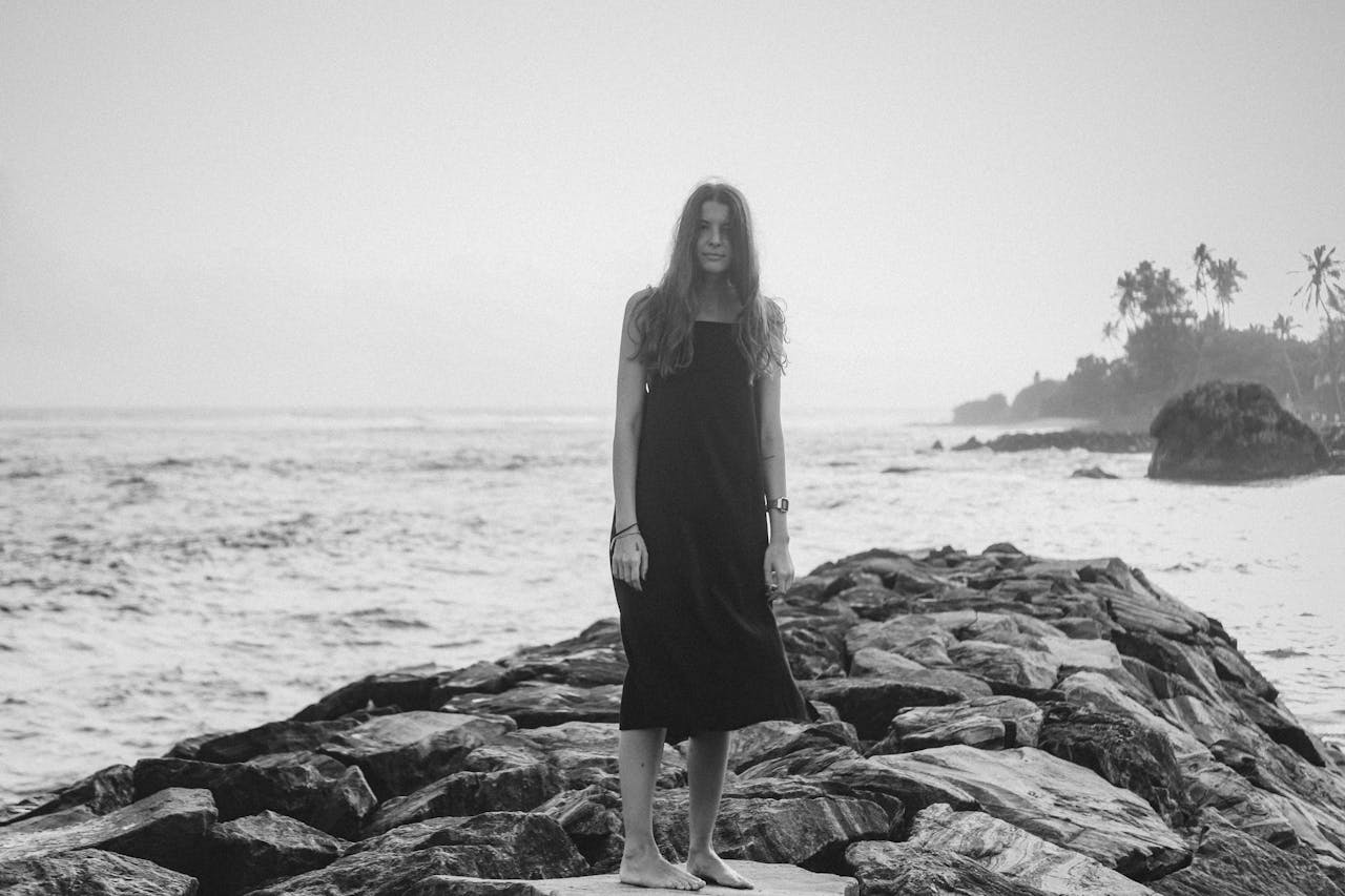 A woman in a black dress stands on a rocky shoreline, facing the sea, in a serene black and white scene.